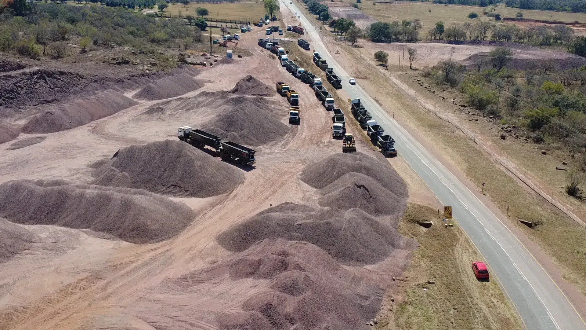 Drone view of trucks queued beside material stockpiles next to a live road under construction